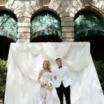 Bride in textured white ballgown and groom in white tuxedo jacket pose beneath white ceremony draping