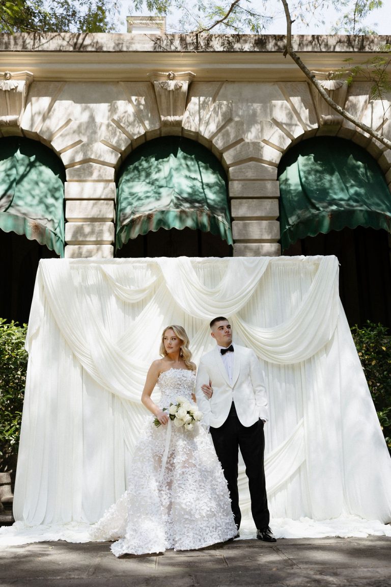 Bride in textured white ballgown and groom in white tuxedo jacket pose beneath white ceremony draping