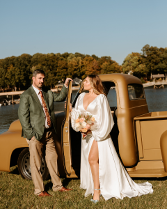 The couple standing beside a vintage truck by the water, the bride holding a bouquet and wearing a flowing white gown while the groom wears a green suit.