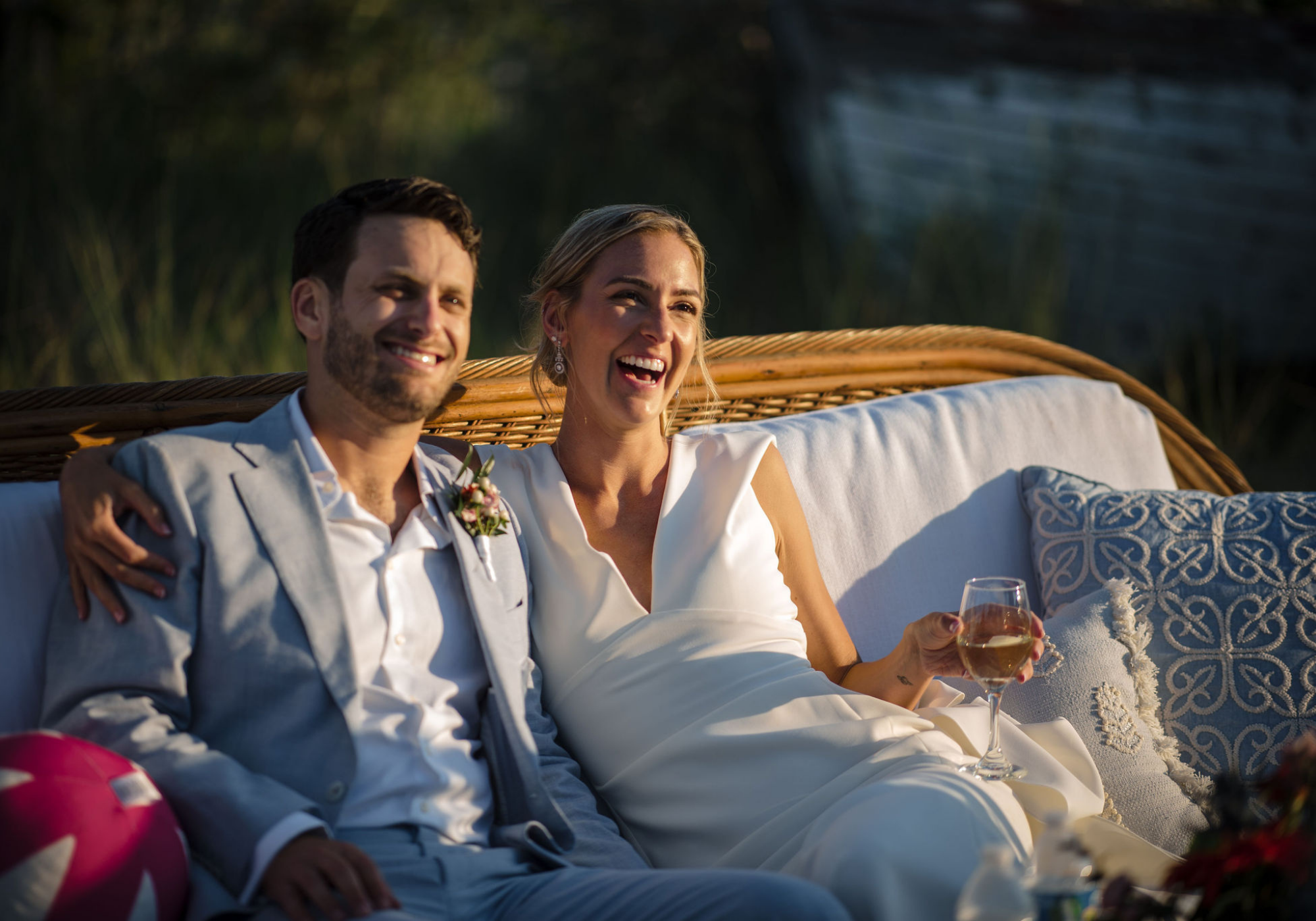 Bride and groom relax together in a vintage wooden boat beside the water, paired with close-up details of their coastal reception décor, including colorful florals, blue linens, woven textures, and candlelight.