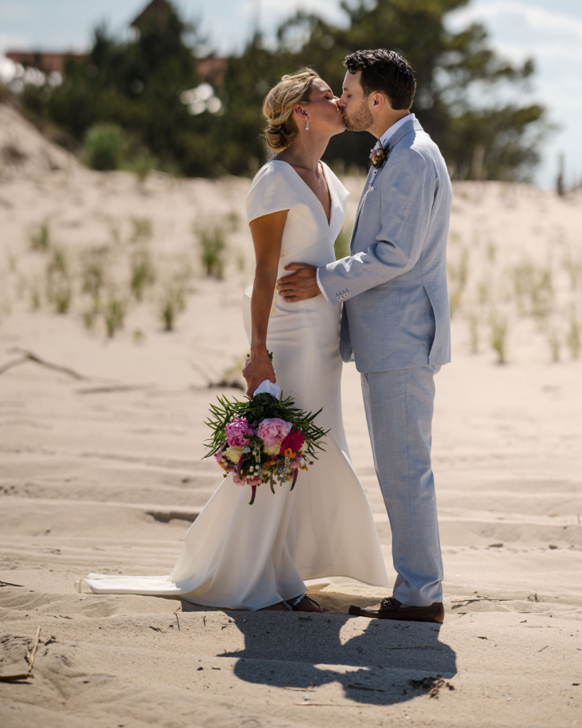 Bride and groom share a kiss on the beach with the ocean behind them during their Delaware coastal wedding portraits.
