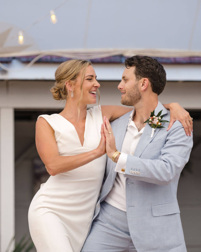 Bride and groom smile at one another while holding hands on the beach before their Delaware wedding ceremony.