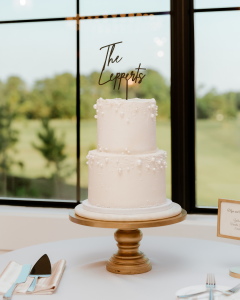 Two-tier white buttercream wedding cake with delicate pearl detailing and “The Lepperts” topper by Mill Stream Farm BakeStudio, displayed on a gold stand at a Delaware wedding reception.