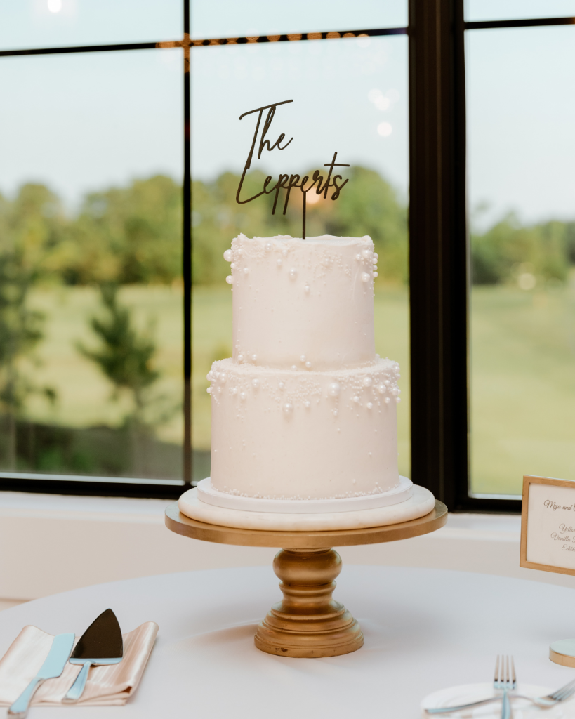 Two-tier white buttercream wedding cake with delicate pearl detailing and “The Lepperts” topper by Mill Stream Farm BakeStudio, displayed on a gold stand at a Delaware wedding reception.
