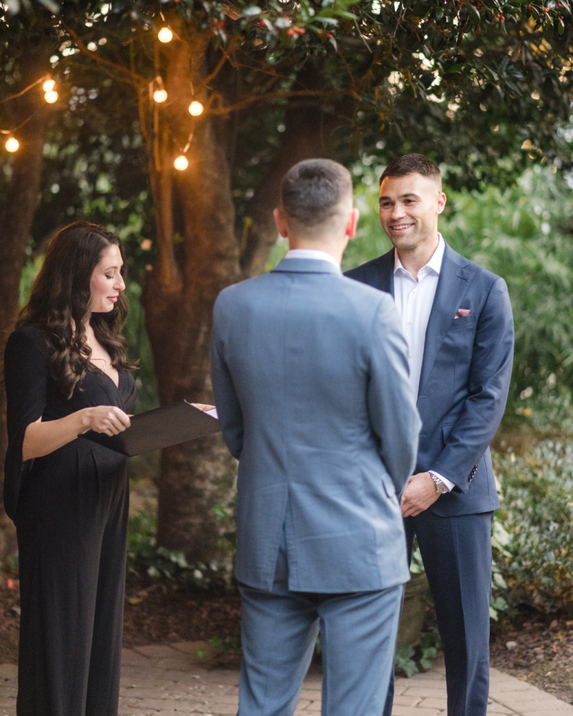 A wedding officiant stands with two grooms as they exchange vows during a romantic outdoor ceremony with warm string lights.