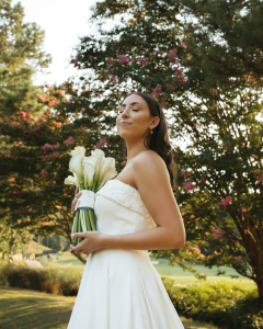 Bride posing outdoors with bouquet of white calla lilies, surrounded by greenery.