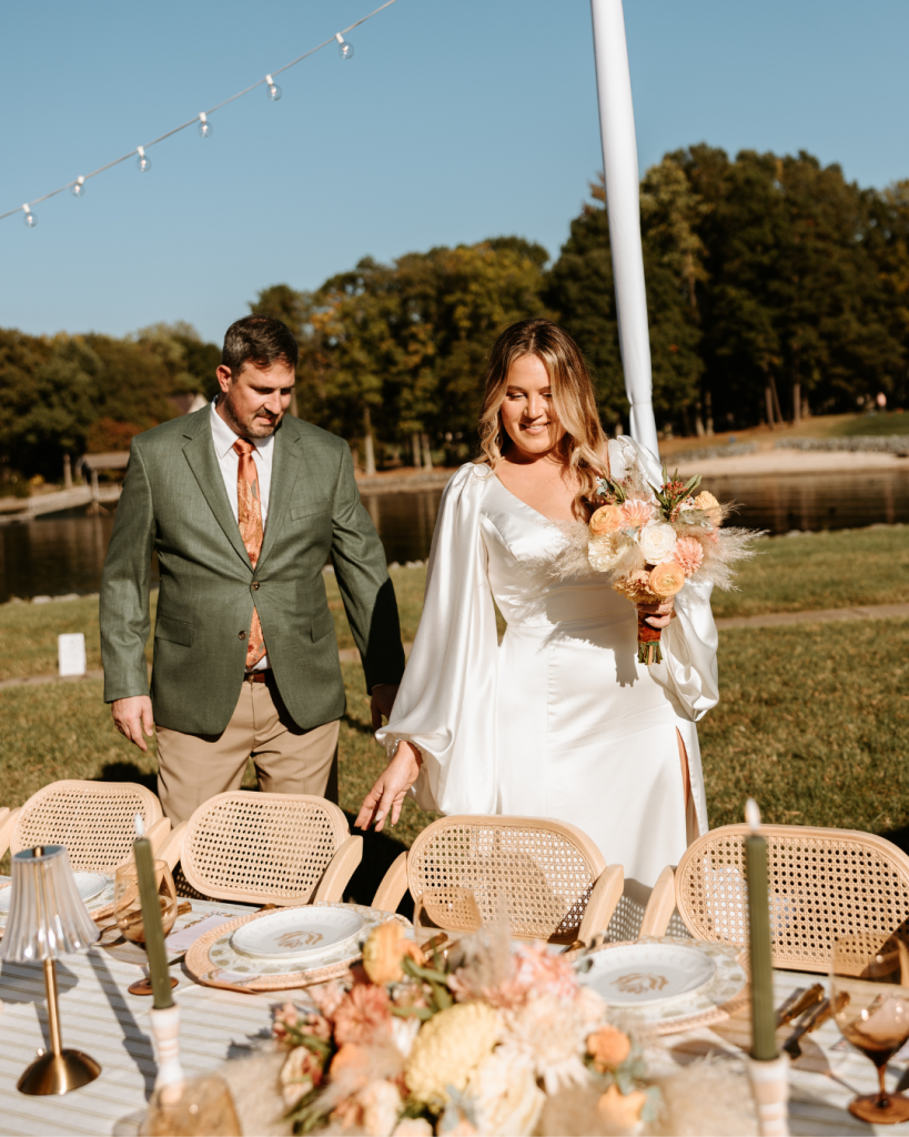 The couple walking hand in hand past a beautifully styled reception table outdoors by the water.