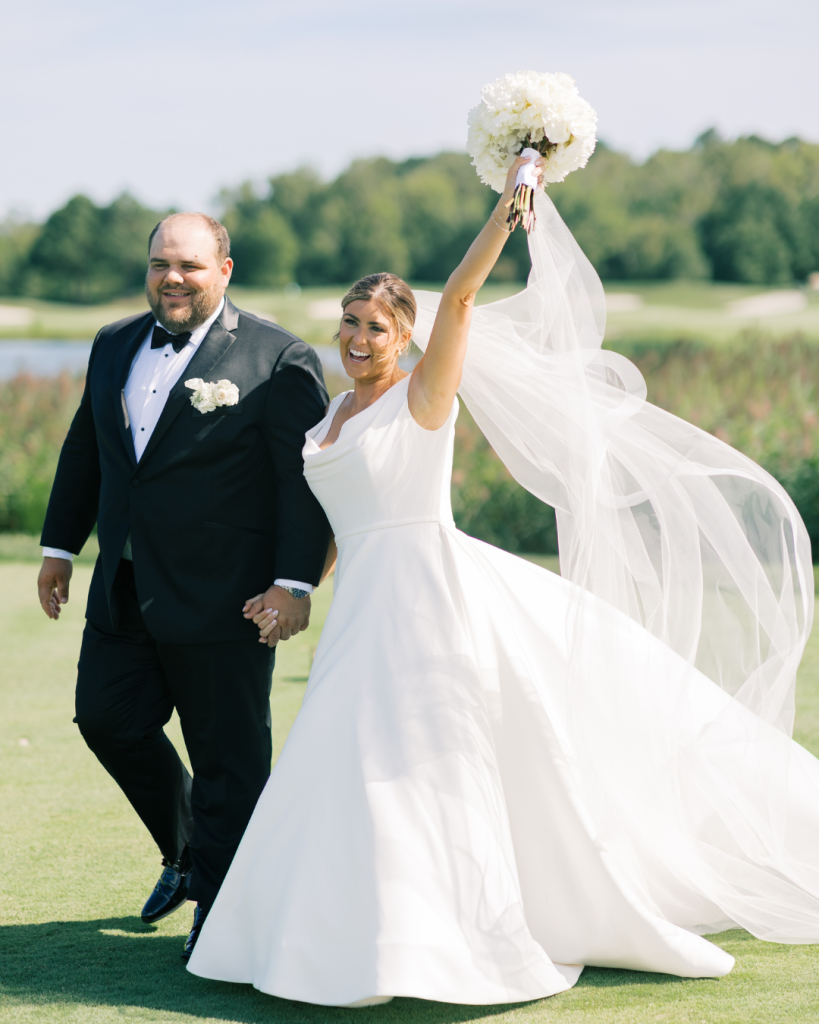 Bride and groom walking together on golf course lawn with veil flowing during coastal wedding at Bayside Resort Golf Club in Selbyville, Delaware.