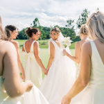 Bride walking with bridesmaids on golf course lawn holding white bouquets during coastal Delaware wedding at Bayside Resort Golf Club.
