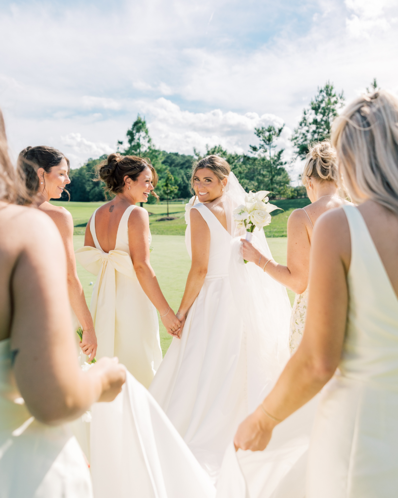 Bride walking with bridesmaids on golf course lawn holding white bouquets during coastal Delaware wedding at Bayside Resort Golf Club.