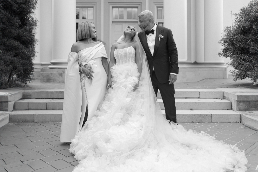 A bride in a voluminous ruffled gown stands between two adults, one in a formal dress and the other in a tuxedo, on a stone pathway outside a building with columns.