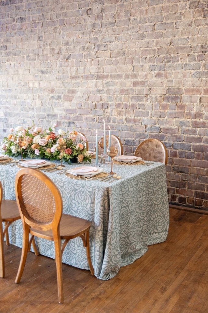 Elegant event rental tablescape in Cleveland featuring a sage patterned tablecloth, cane-back chairs, candles, and peach floral centerpiece against a rustic brick wall.