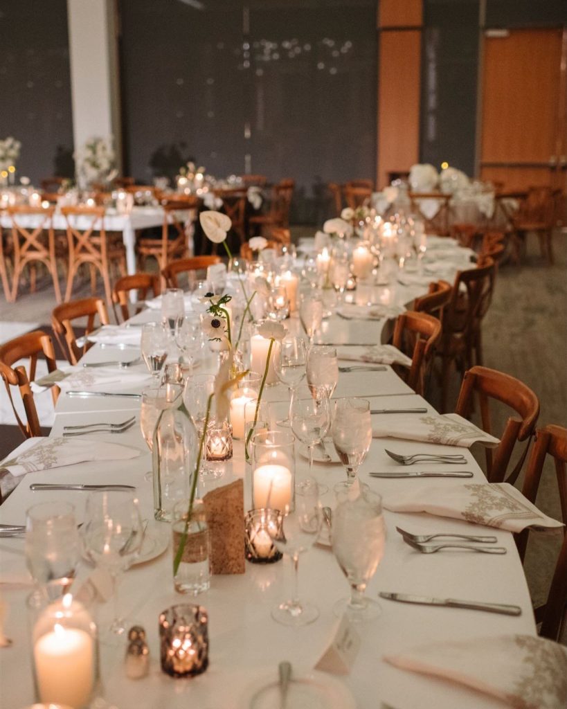 A long wedding reception table decorated with numerous glowing candles, white flowers by Roses + Mint, and elegant place settings.