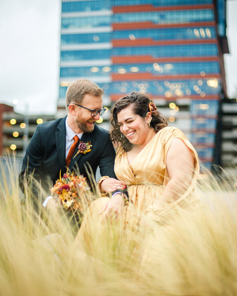 bride and groom in a grass field with a city view behind them smiling and laughing