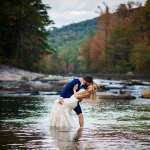 groom dipping his bride in shalow water with trees in the background full of fall colors