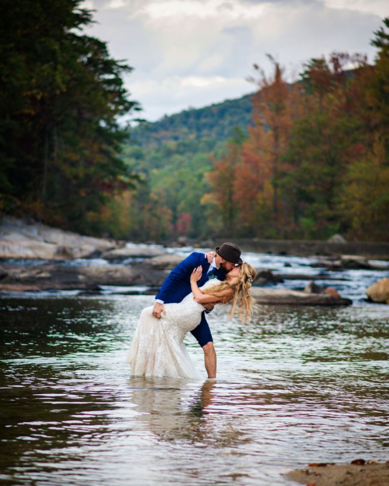 groom dipping his bride in shalow water with trees in the background full of fall colors