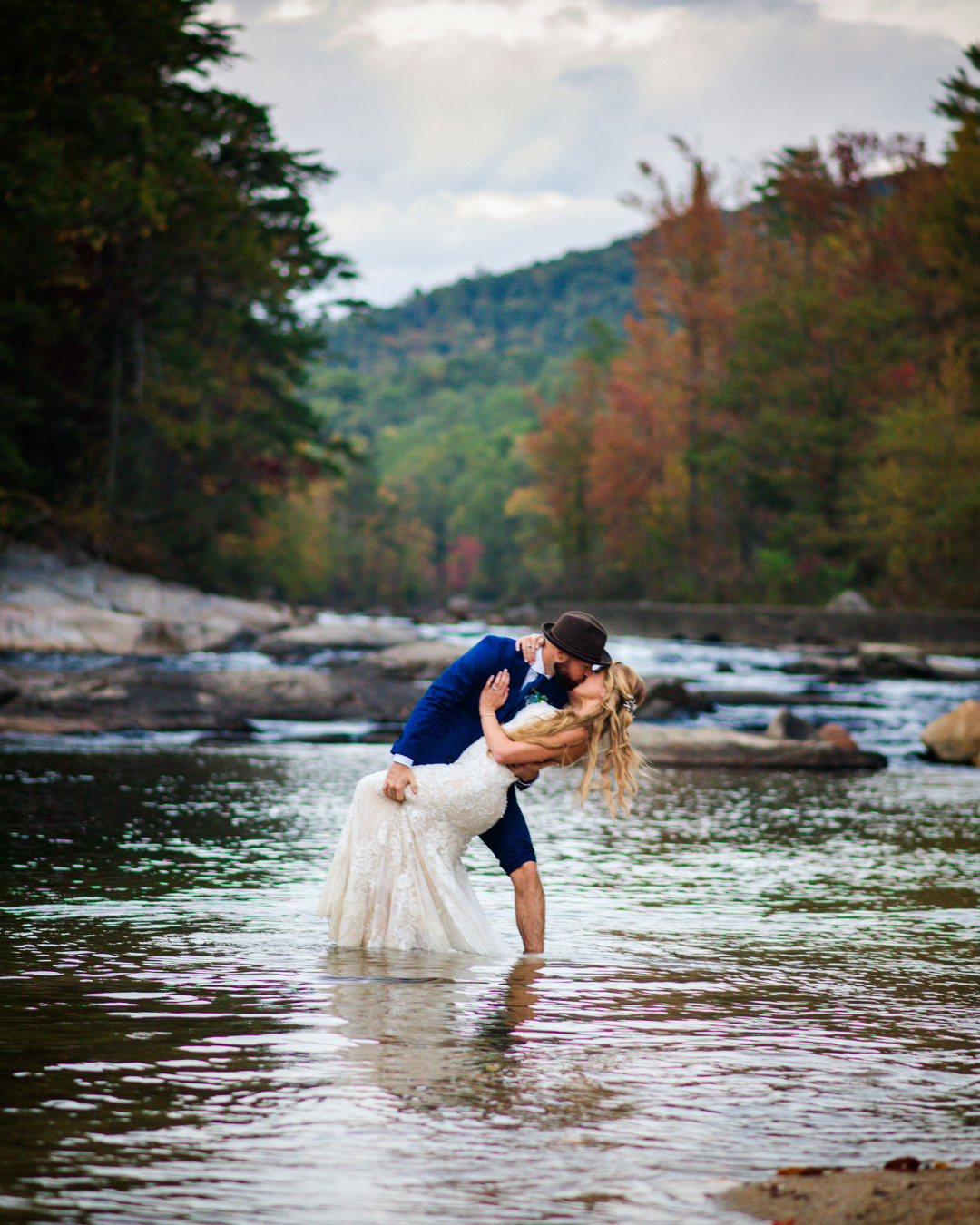 groom dipping his bride in shalow water with trees in the background full of fall colors