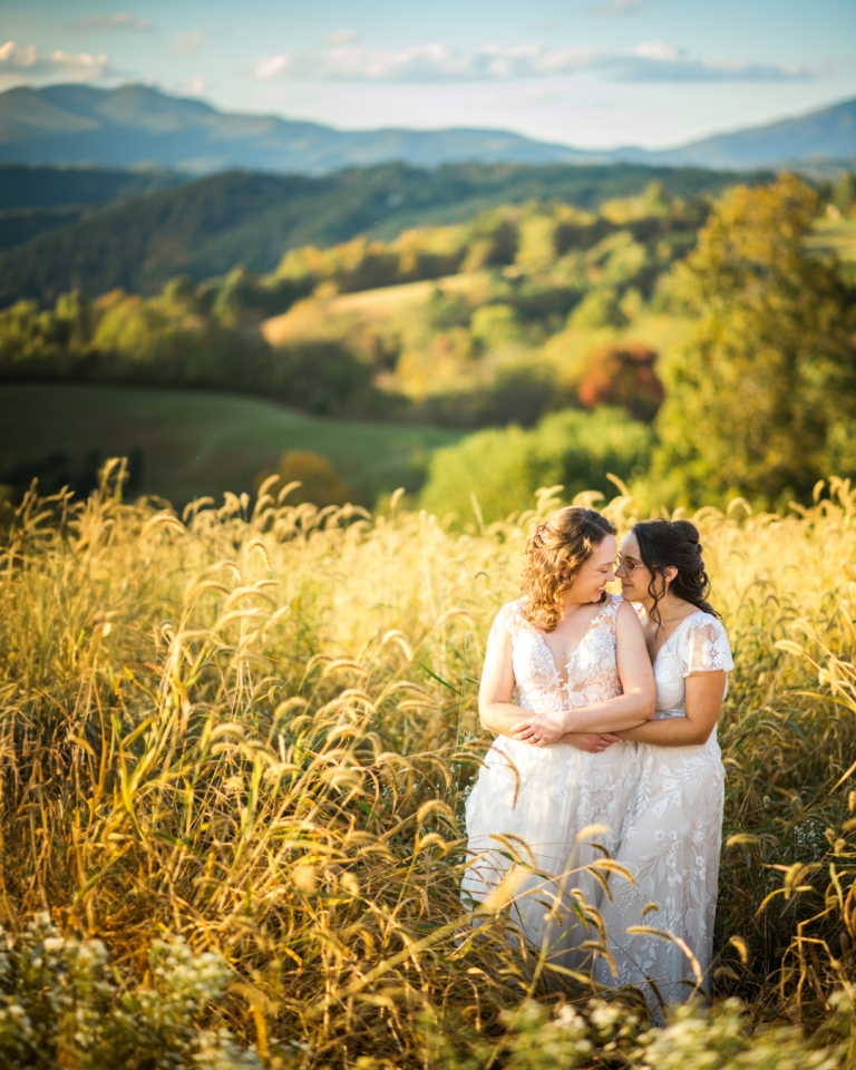 LGBTQ+ couple looking into eachothers eyes in their white bridal dresses with a beautiful mountain view behind them