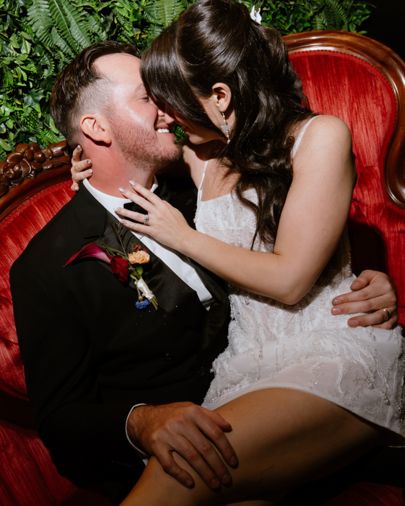 Bride and groom sharing an intimate kiss while seated in a red velvet chair surrounded by greenery.