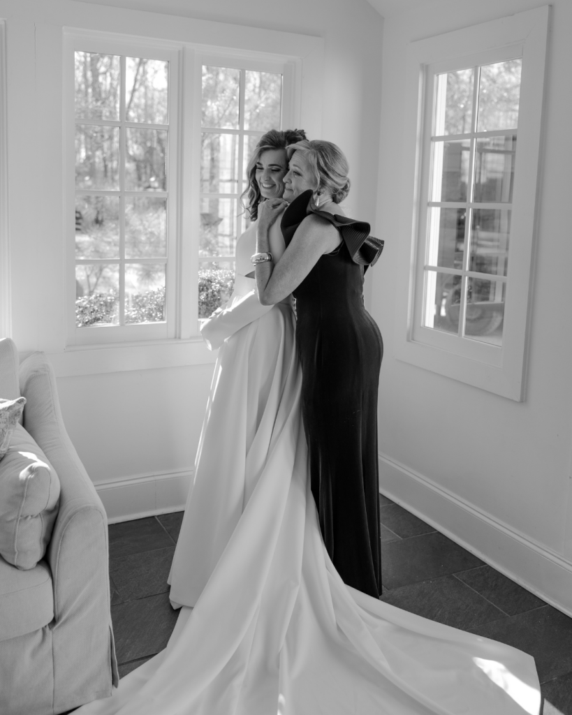 Black and white photo of bride hugging a loved one, showcasing an emotional moment and the elegant silhouette of her gown