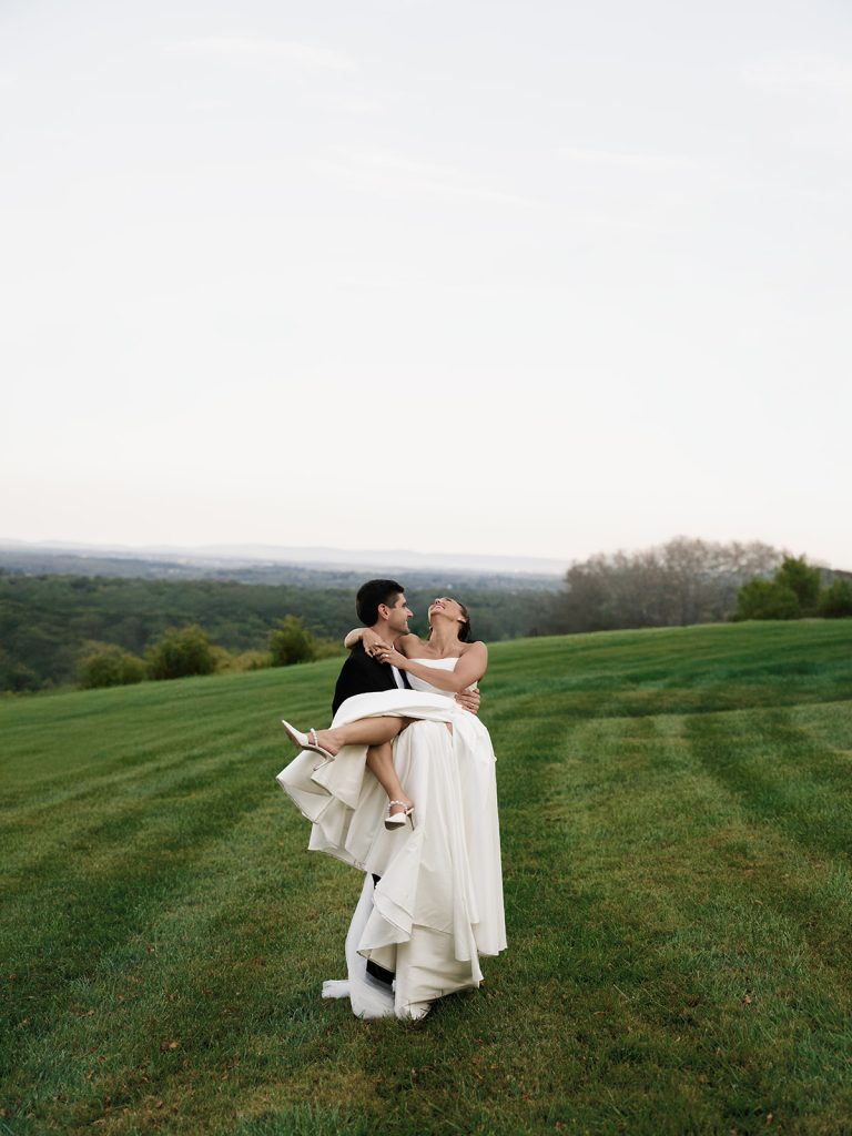 Husband holding wife in arms in green field at wedding venue