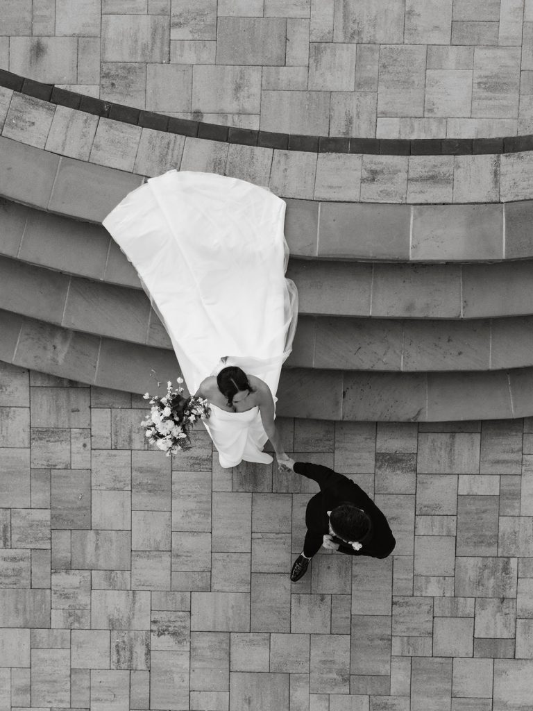 Black and White photo overview of groom and bride walking together down stairs with bridal train trailing behind