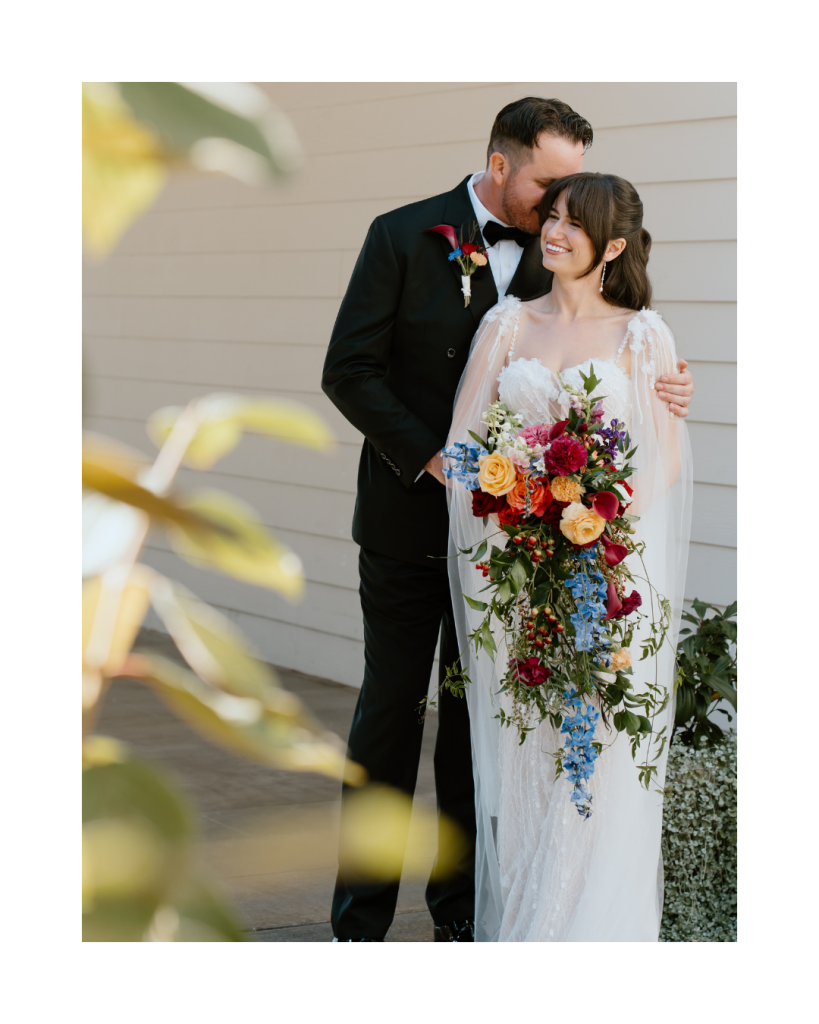 Bride and groom embracing, with the bride holding a cascading bouquet filled with bold, colorful blooms.