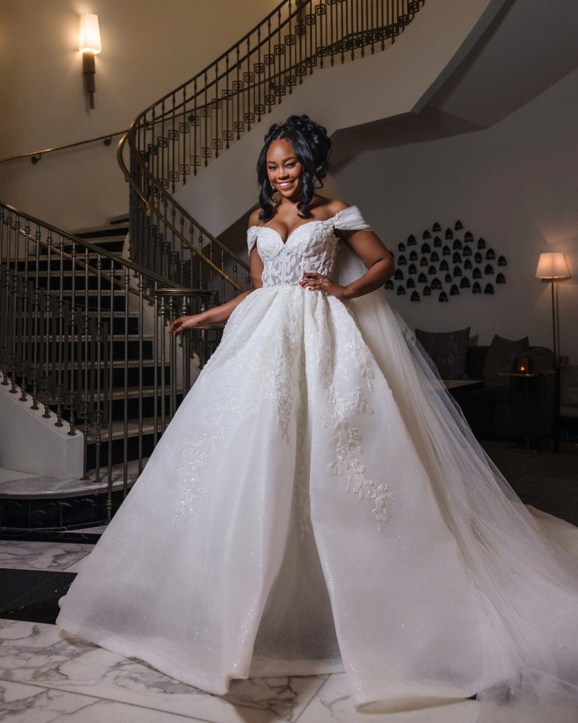 A bride smiles in a voluminous white off-the-shoulder wedding dress with lace detailing, standing on a marble floor in front of a grand staircase.