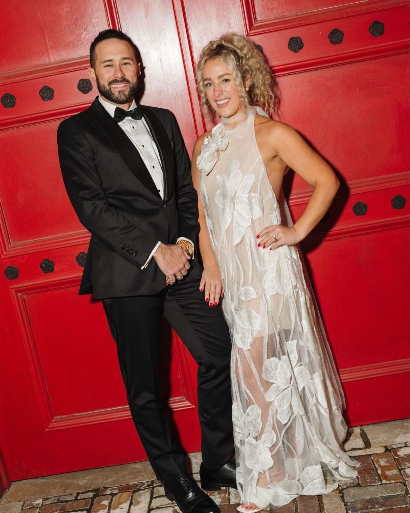 A bride in a sheer floral reception dress and a groom in a tuxedo pose together in front of a large red door.