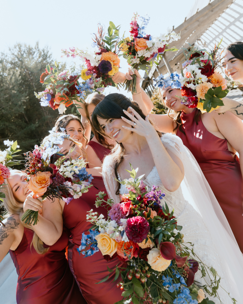 Bride laughing with bridesmaids in coordinated dresses, all holding bright, lush bouquets in an outdoor setting.