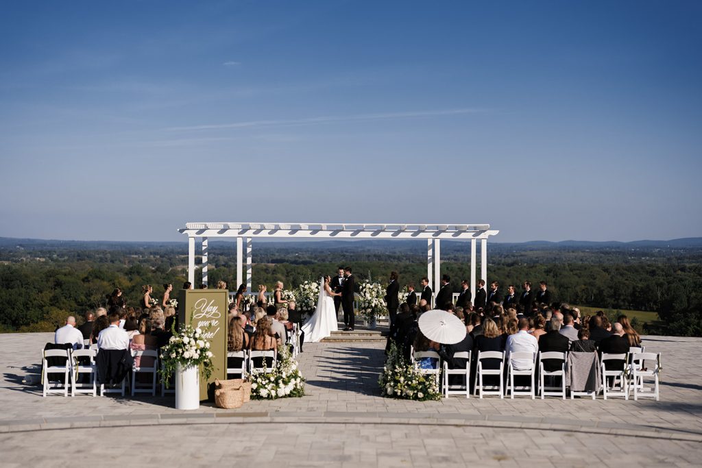 overview photo of wedding ceremony overlooking scenic mountains. Guests are holding white parasols featuring green welcome sign