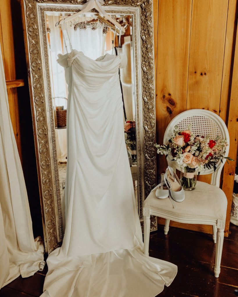 Draped satin wedding gown hanging beside vintage mirror and bridal bouquet inside rustic Delaware farm venue