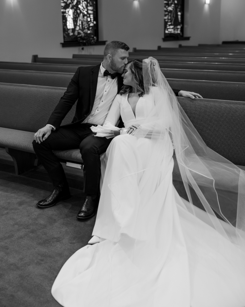 Black and white image of bride and groom sitting together in a church pew, sharing a quiet and intimate moment