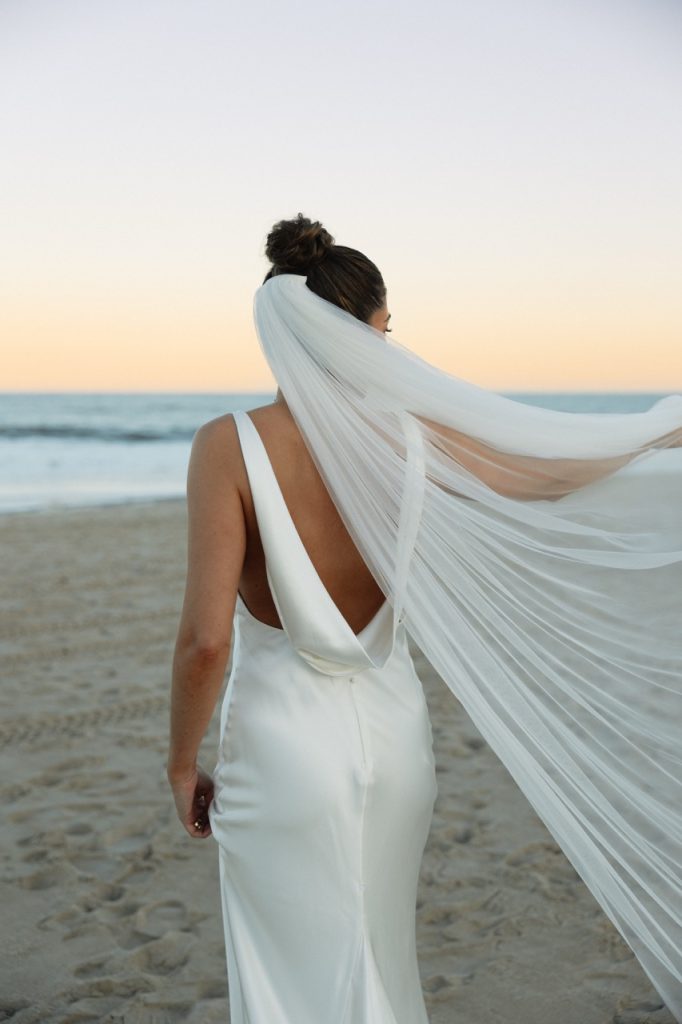 Bride walking along shoreline with veil flowing in ocean breeze during sunset portraits at Addy Sea wedding in Bethany Beach, Delaware.