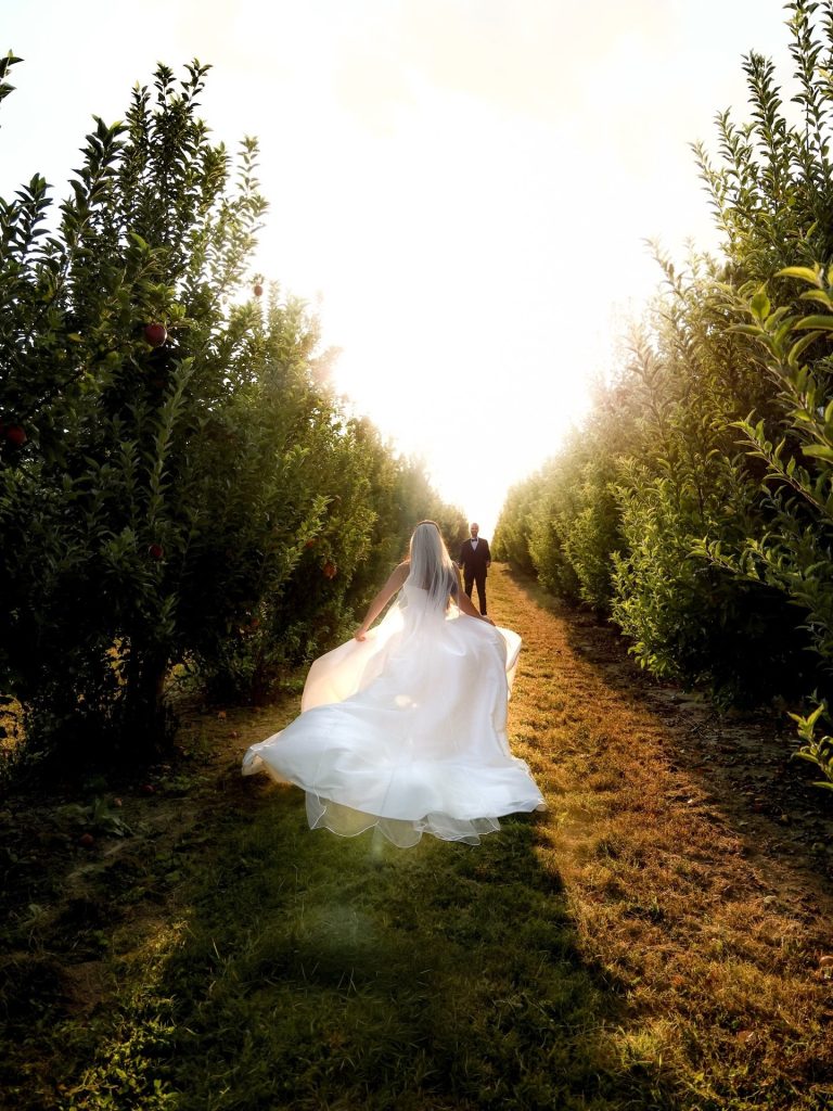 A bride in a Mia Grace Bridal gown runs through a sunlit apple orchard toward her groom.
