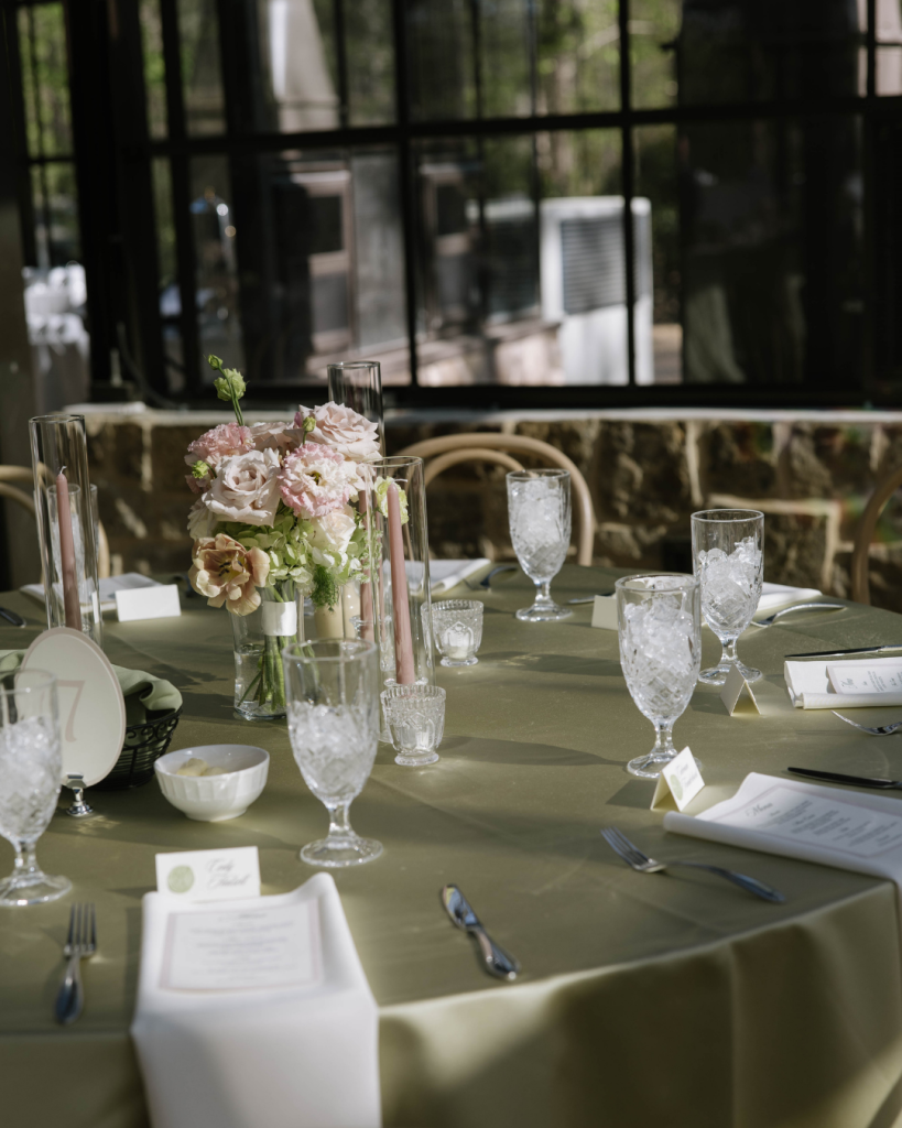 Reception table with green linens, crystal glassware, and floral centerpiece near large windows