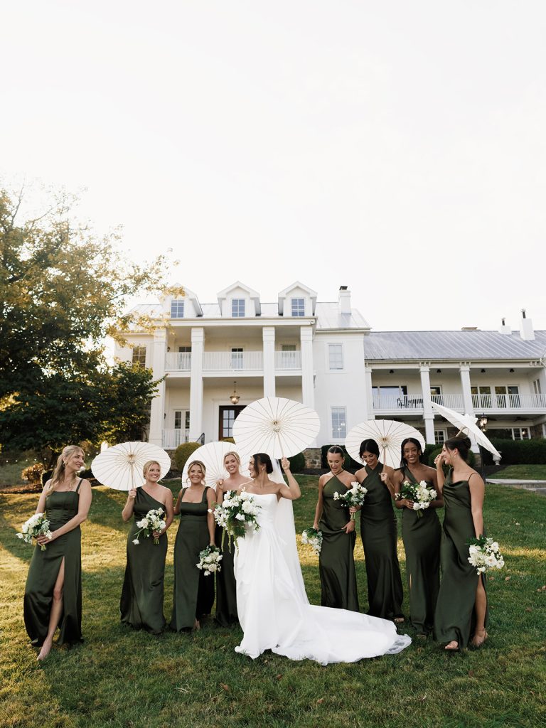 Bridesmaid and bride pose in front of mansion style wedding venue on green lawn