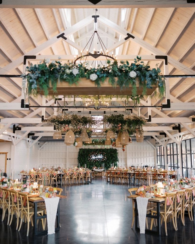 A spacious wedding reception area indoors at Meadows at Mossy Creek with long tables set for dining, adorned with floral arrangements and hanging greenery. A large floral wall serves as a backdrop for the head table.