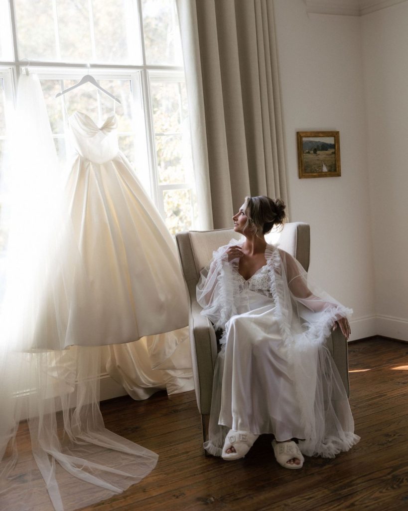 A wedding dress hangs on a hanger by a window, while a person in a white robe and slippers sits in a chair nearby.