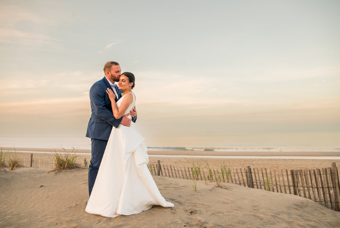 Bride and groom posing on the sand at Addy Sea Historic Oceanfront Inn in Bethany Beach, Delaware, with ocean waves and dunes behind them
