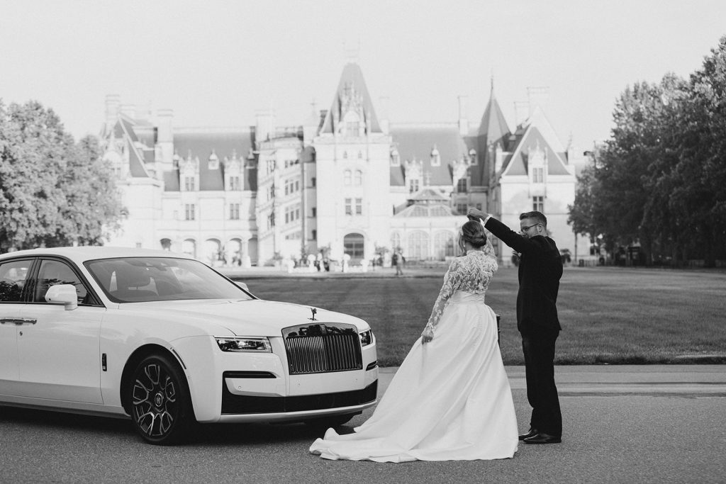 groom spinning his bride next to a white classic getaway car in front of the biltmore estate in north carolina