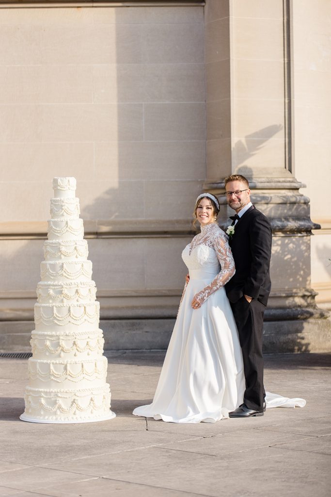 bride and groom standing next to a 6 ft tall white wedding cake