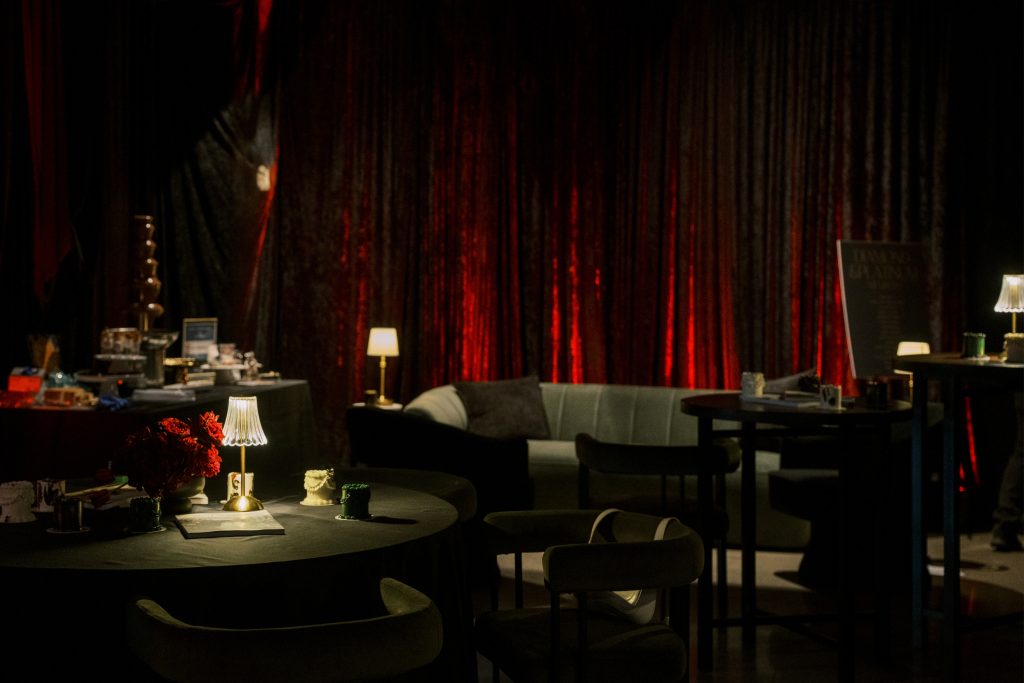 Elegant event setup featuring a dark table with flowers, a lamp, and a backdrop of rich red drapery, creating a sophisticated atmosphere.