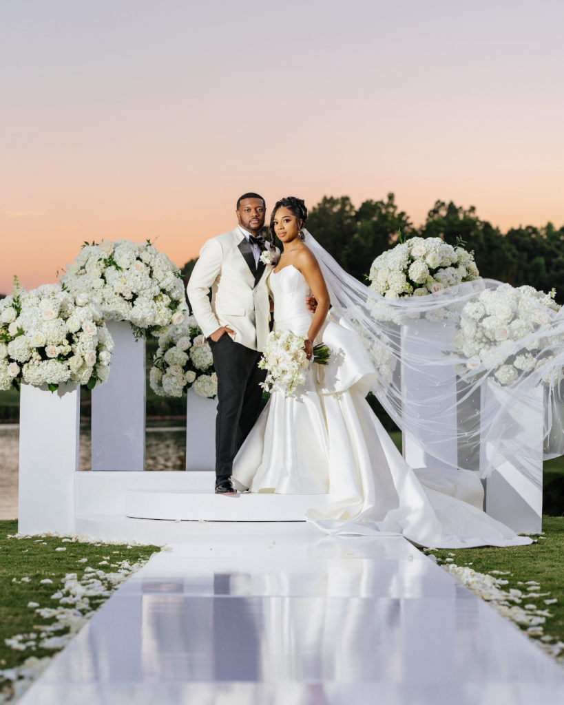 Luxury wedding ceremony at Trump National Golf Course Charlotte featuring a bride and groom standing on a white stage surrounded by lush white floral arrangements and a reflective aisle