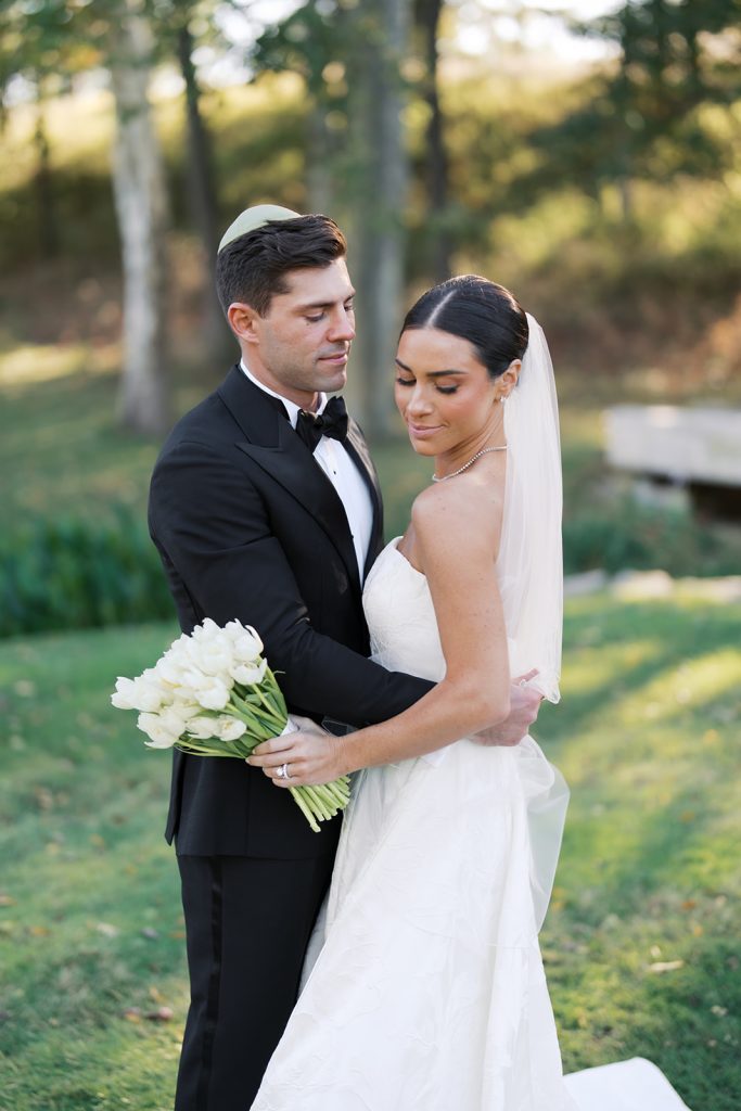 The groom embraces the bride, who wears a strapless gown from Mia Grace Bridal and holds a white tulip bouquet.