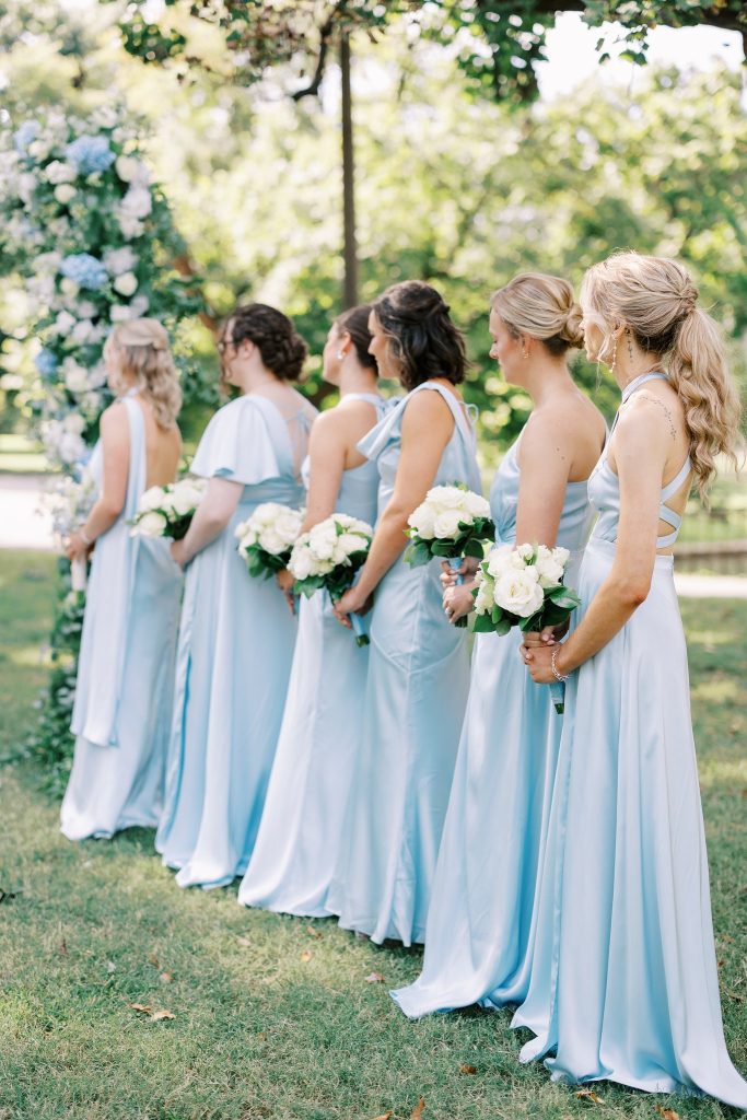 Six bridesmaids dressed in matching light blue satin gowns stand in a row outdoors holding bouquets of white roses at a wedding planned by Hawthorne Creative Co.