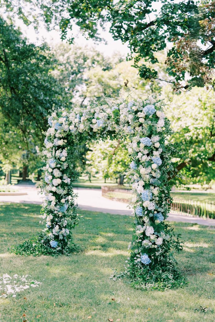 A rectangular wedding arch lushly covered in white roses, blue hydrangeas, and green foliage stands on a grassy lawn surrounded by trees for a wedding planned by Hawthorne Creative Co.