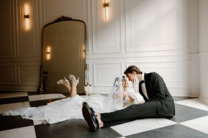 A bride and groom sitting on a black and white checkered floor, facing each other intimately, with the bride's legs and wedding dress fanned out behind her.