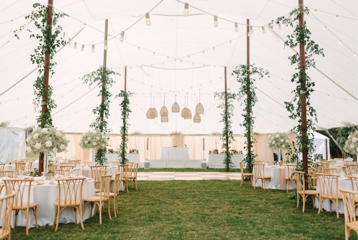 Interior of a sailcloth wedding tent on the Eastern Shore with long tables, wood chairs, greenery-wrapped poles, and café lighting overhead