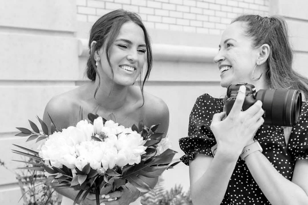 A smiling bride holding a bouquet of white flowers while Katrina Poggio, the wedding photographer, shows her an image on her camera.
